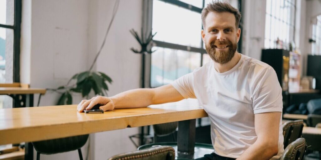 Jan Hicl Sitz am tisch im Büro, bärtigerjunger Mann weißer T-Shirt. freundliches Lächerln
