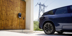 BMW bidirektionales Laden. BMW vor Scheune, Strommast blauer Himmel Lade-Wallbox.