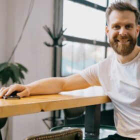 Jan Hicl Sitz am tisch im Büro, bärtigerjunger Mann weißer T-Shirt. freundliches Lächerln